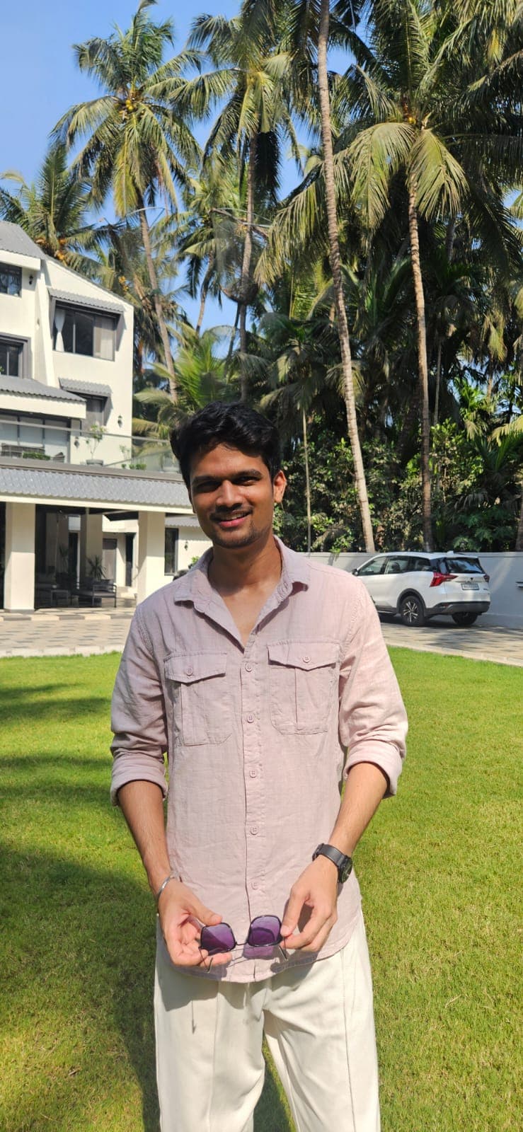 Anup Nagdeve smiling outdoors in front of palm trees, holding a pair of sunglasses
